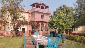 A dining area with a table set for six, surrounded by blue chairs with brown cushions, in front of a pink building with arches and a tower. - Heritage Lotwara Fort (Baswa)