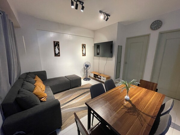 Un salón moderno con un sofá de color oscuro, una mesa de comedor de madera y un televisor de pantalla plana montado en la pared.