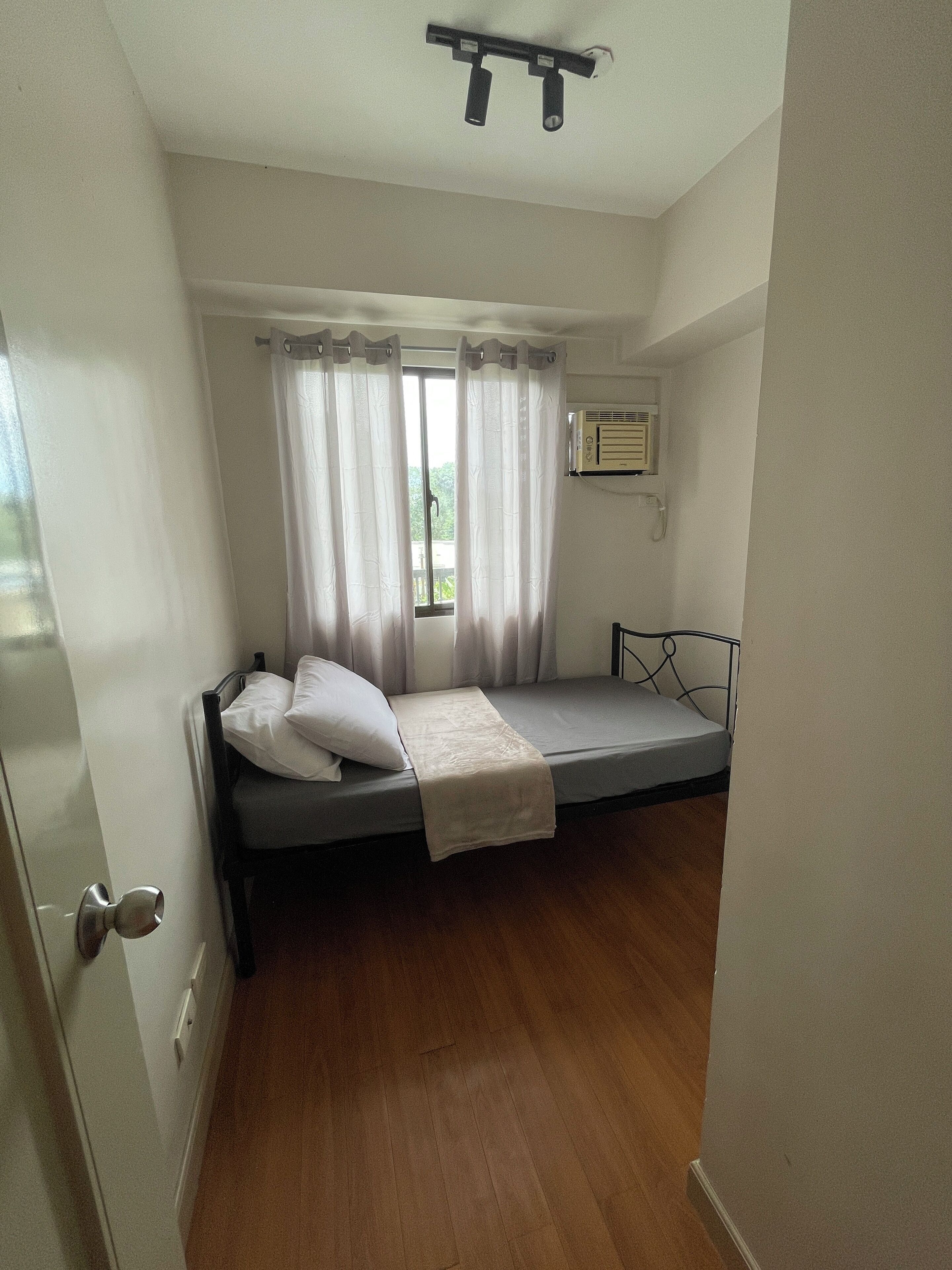 A small, single-bed room with a wooden floor, a bed with a white blanket and pillows, a window with curtains, and a wall-mounted air conditioning unit.