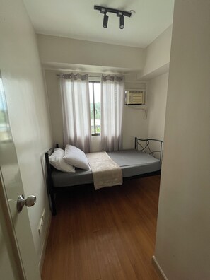 Una habitación pequeña con cama individual, suelo de madera, cama con manta y almohadas blancas, ventana con cortinas y un equipo de aire acondicionado montado en la pared.