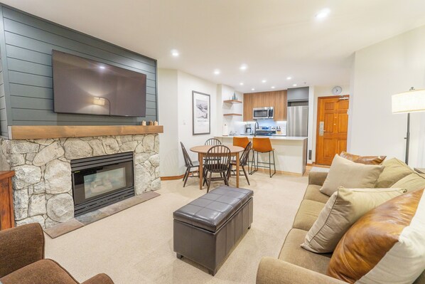 A modern living room with a stone fireplace, a flat-screen TV mounted above it, a dining area with a table and chairs, and a kitchen in the background. - Aspens #218 Prime Ski-in Ski-out! Pool, Hot tubs! (Whistler)
