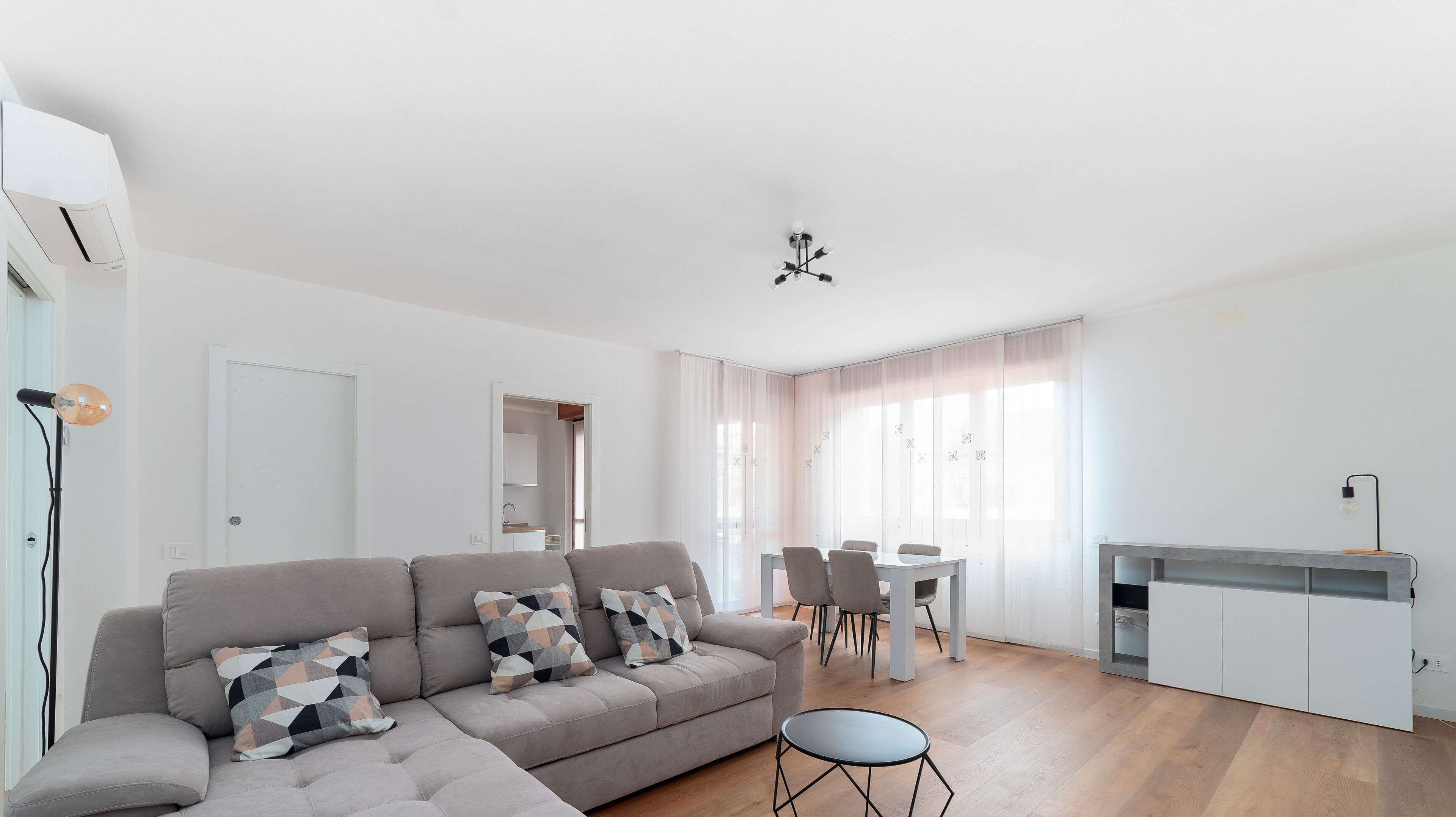 A modern living room with a grey sectional sofa, a small round table, and a dining area with chairs.
