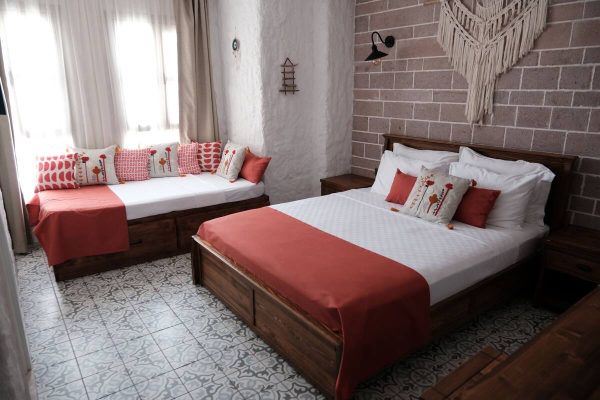 Una habitación con dos camas, cada una con estructura de madera y ropa de cama blanca. El suelo tiene baldosas estampadas y una pared de ladrillo decorada con un tapiz de macramé.