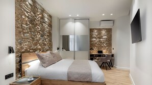 A modern bedroom with a stone wall, a bed with a wooden frame, a desk with a chair, and a TV mounted on the wall. - J&A Luxury Residence (Athens)