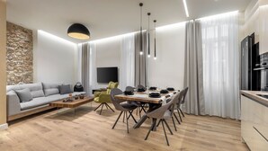 A modern living room with a sofa, coffee table, dining table, and chairs. There is a stone wall, a flat-screen TV, and a kitchen area with a refrigerator. - J&A Luxury Residence (Athens)