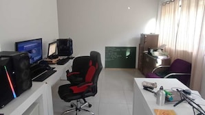Ein kleiner Büroraum mit zwei Schreibtischen, Computern, einem schwarzen Bürostuhl, einer Tafel und einem Fenster mit Vorhängen.