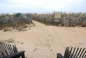 Un sentier de sable qui longe la plage et mène à un panneau indiquant le passage d’une dune, le tout étant bordé de clôtures en bois et d’une végétation clairsemée.