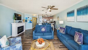 A living room with a fireplace, blue sofas, a coffee table with a floral arrangement, and a kitchen in the background. - Includes Xplorie Activities & Linens. PYRAMID-2BR OFcondo w/outdoor pool. (Ocean City)