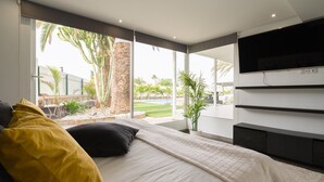 A modern bedroom with a large bed, a TV mounted on the wall, and a view of a pool and palm trees outside. - Villa Luz de Maspalomas (Maspalomas)