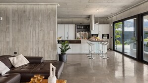 A modern living room with a dark leather sofa, a wooden coffee table, and a large window offering a view of the kitchen and outdoor area.