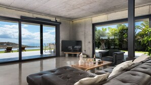 A modern living room with a large leather sofa, a wooden coffee table with glasses and a bottle, and a TV. The room has large glass doors leading to an outdoor area with a pool and greenery.