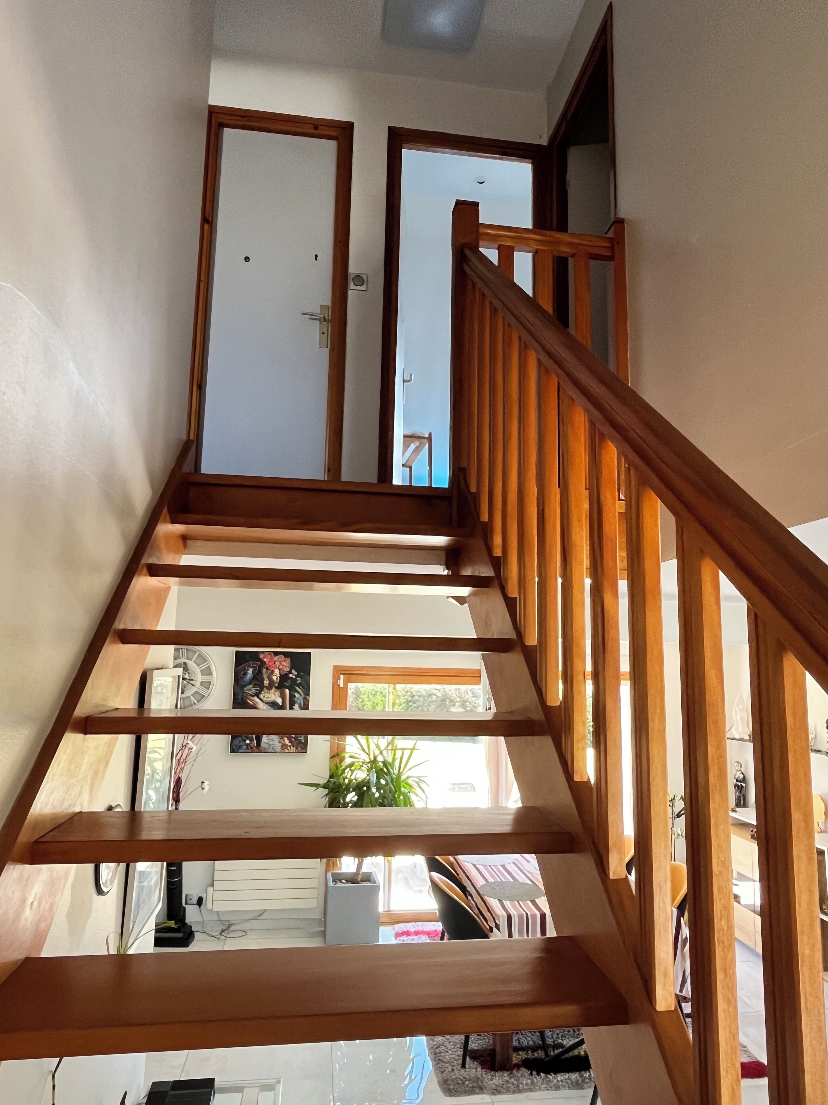 Un escalier en bois offrant une vue sur une pièce située en contrebas, où l’on peut apercevoir une porte, une horloge et une plante.