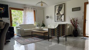 A modern living room with a white sofa, a coffee table, and a patterned chair. There is a framed artwork on the wall and a potted plant on the floor.