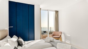 Een moderne hotelkamer met een groot bed, een blauwe accentmuur, uitzicht op het groen en een zithoek met een stoel en een tafel.