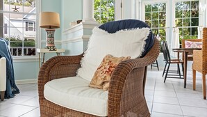 Una silla de mimbre con un cojín blanco y un almohadón decorativo, ubicada cerca de una ventana con un adorno de estrella de mar.