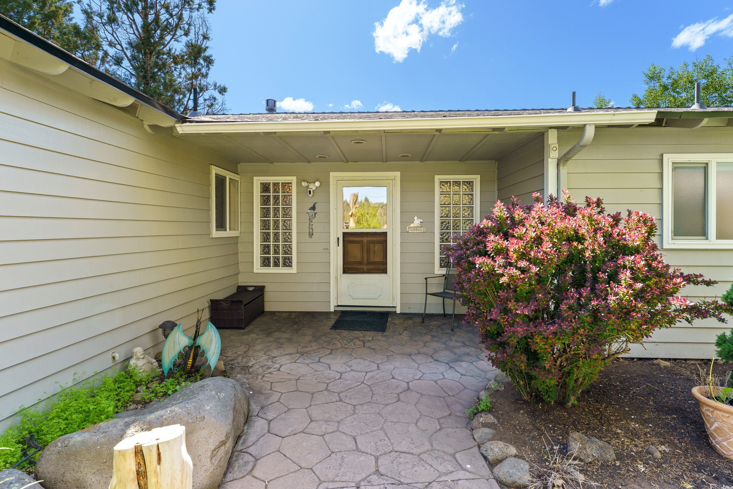 A house with a stone patio, a front door, and a small garden with pink flowers.