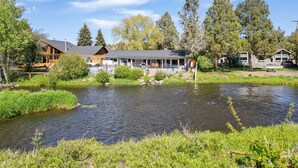 A lakeside cabin with a deck overlooking the water, surrounded by trees and a clear sky. - Otter Run - Riverfront Paradise in Tumalo, Hot Tub, Sauna, Deck, Pet Friendly! (Tumalo)