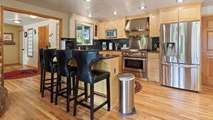 A modern kitchen with wooden cabinets, stainless steel appliances, and a bar area with stools. - Otter Run - Riverfront Paradise in Tumalo, Hot Tub, Sauna, Deck, Pet Friendly! (Tumalo)