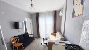 Un salon moderne comprenant un canapé, une chaise en bois, une petite table, un téléviseur fixé au mur et un coin cuisine avec une cuisinière.