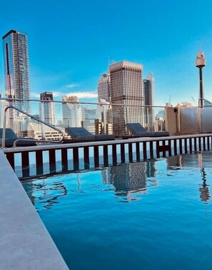 Una piscina en la azotea con sillones de descanso, rodeada de edificios altos.