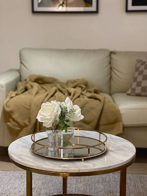 Un tavolo rotondo con cornice dorata e piano in vetro, posizionato di fronte a un divano beige con una coperta marrone.