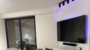 Ein modernes Wohnzimmer mit Flachbildfernseher an einer weißen Wand, zwei grauen Sesseln, einem kleinen weißen Schreibtisch und einer Glastür zum Küchenbereich.