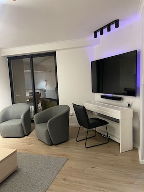 A modern living room with a flat-screen TV mounted on a white wall, two grey armchairs, a small white desk, and a sliding glass door leading to a kitchen area. - Elegant Hyde Park View Retreat with Rooftop Spa Sauna Pool and Gym (Darlinghurst)
