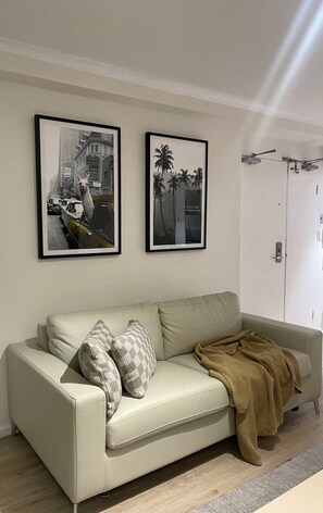 A living room with a beige sofa, two framed pictures on the wall, a door, and a brown throw blanket on the sofa. - Elegant Hyde Park View Retreat with Rooftop Spa Sauna Pool and Gym (Darlinghurst)