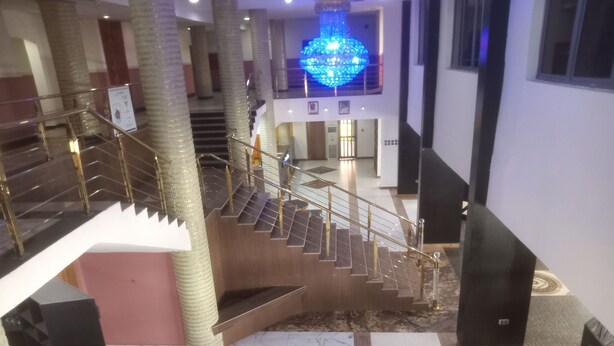Un vestíbulo de hotel con piso de mármol, una escalera con baranda de vidrio y una lámpara decorativa azul.