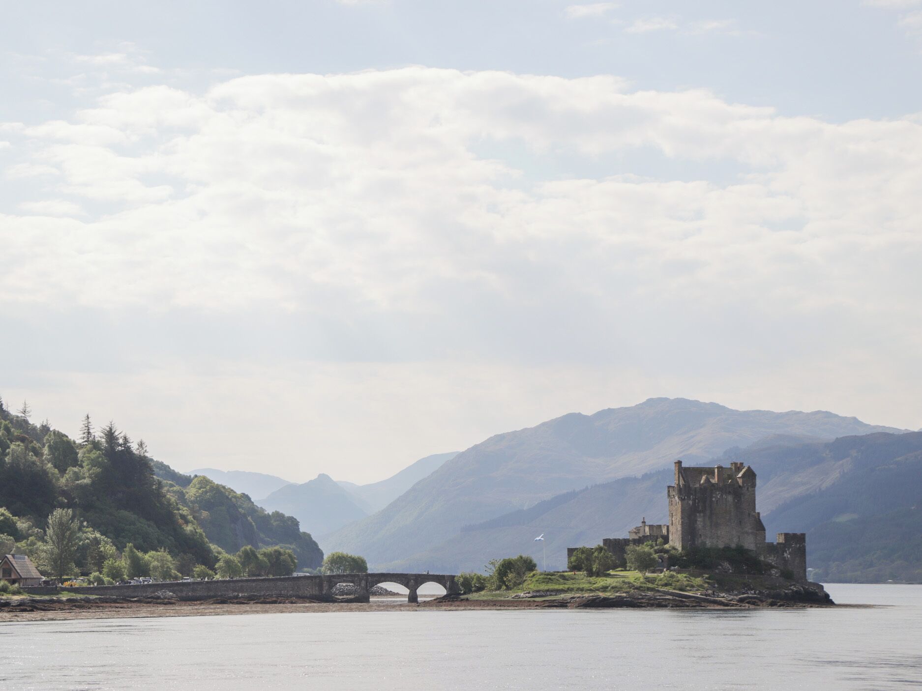A historic castle on a lakeside island, a stone bridge leading to it, and mountains in the background.