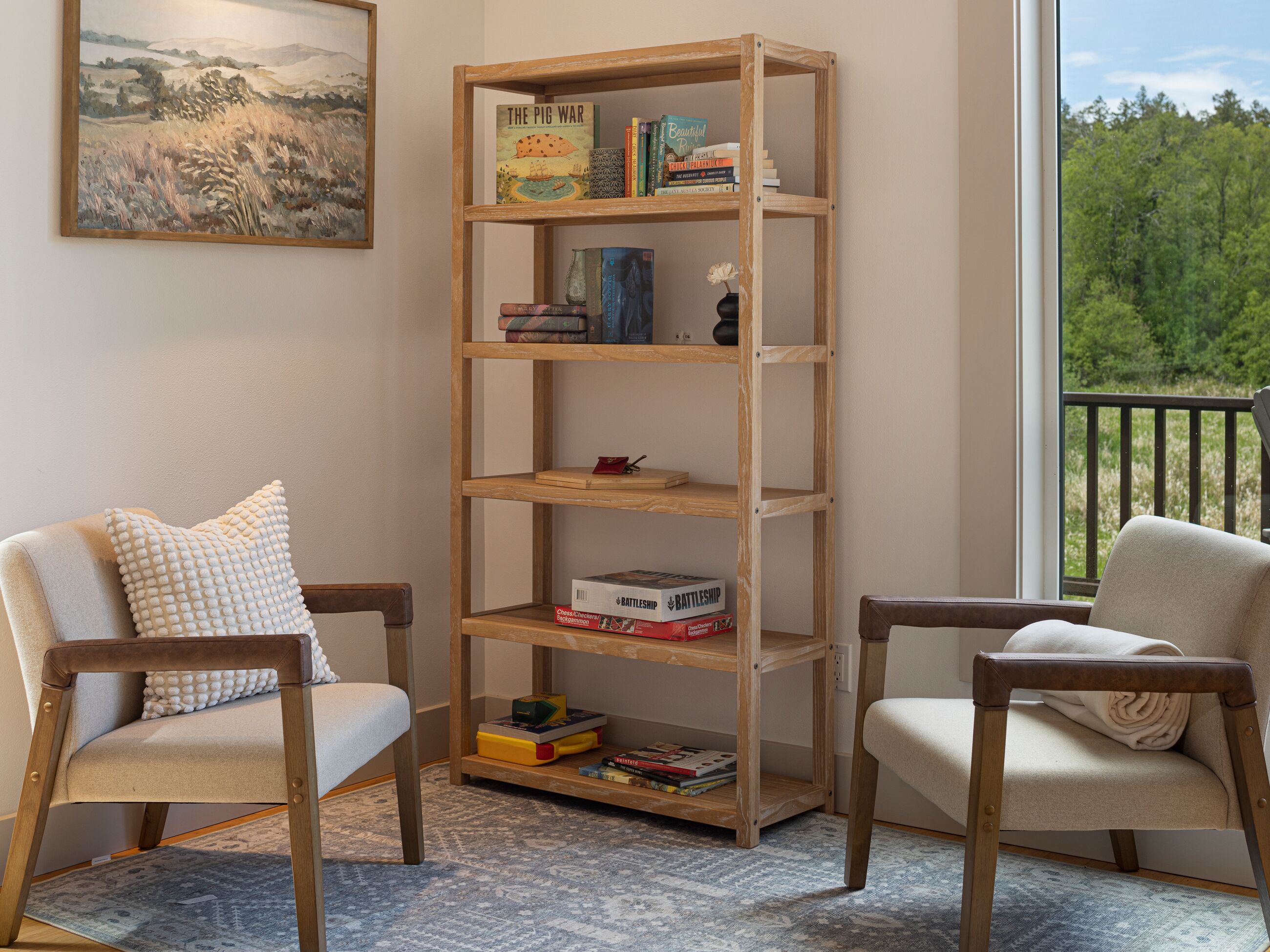 A room with a wooden bookshelf, two armchairs, a painting on the wall, and a view of greenery through a window.