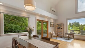 A spacious room with a wooden dining table, chairs, a bookshelf, and a view of greenery through large windows. - Island Escape with Comfort and Privacy -Margo House (Friday Harbor)
