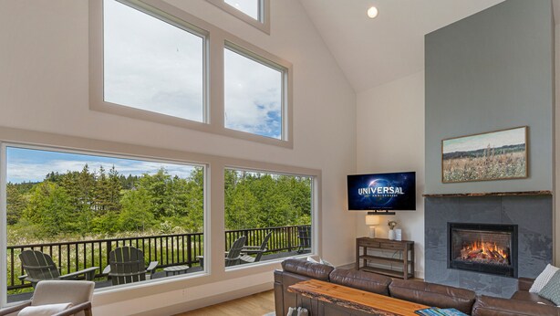 A spacious living room with a large window overlooking a forest, a fireplace, and a television mounted on the wall. - Island Escape with Comfort and Privacy -Margo House (Friday Harbor)