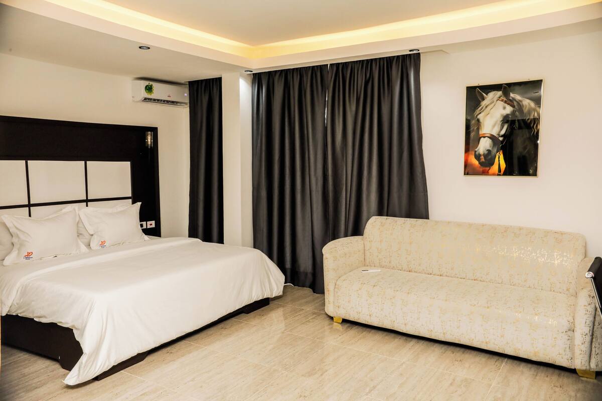 A hotel room with a bed, a sofa, a painting of a horse, and an air conditioning unit.
