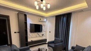 A modern living room with a flat-screen TV, dark sofas, a glass coffee table, and a patterned rug.