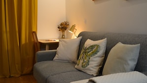 A grey sofa with a white and a butterfly-patterned pillow, a small table with a lamp, and a vase with dried flowers.