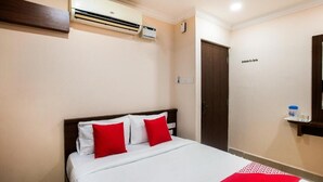 Ein Hotelzimmer mit einem Bett, roten Kissen, einer weißen Bettdecke, einer Klimaanlage, einer Holztür und einem kleinen Regal mit einer Wasserflasche und Gläsern.