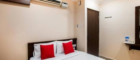 A hotel room with a bed, red pillows, a white bedspread, an air conditioning unit, a wooden door, and a small shelf with a water bottle and glasses.