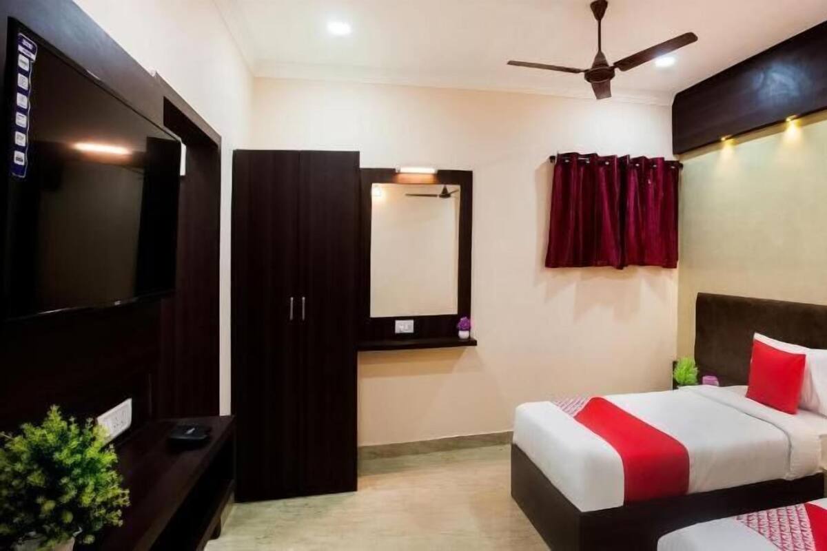 Habitación de hotel con televisión, armario, espejo, dos camas con ropa de cama roja y blanca, ventilador de techo y cortina roja.