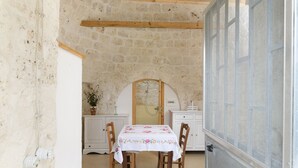 A rustic stone-walled room with a wooden-framed window, a white table with a floral tablecloth, and wooden chairs.
