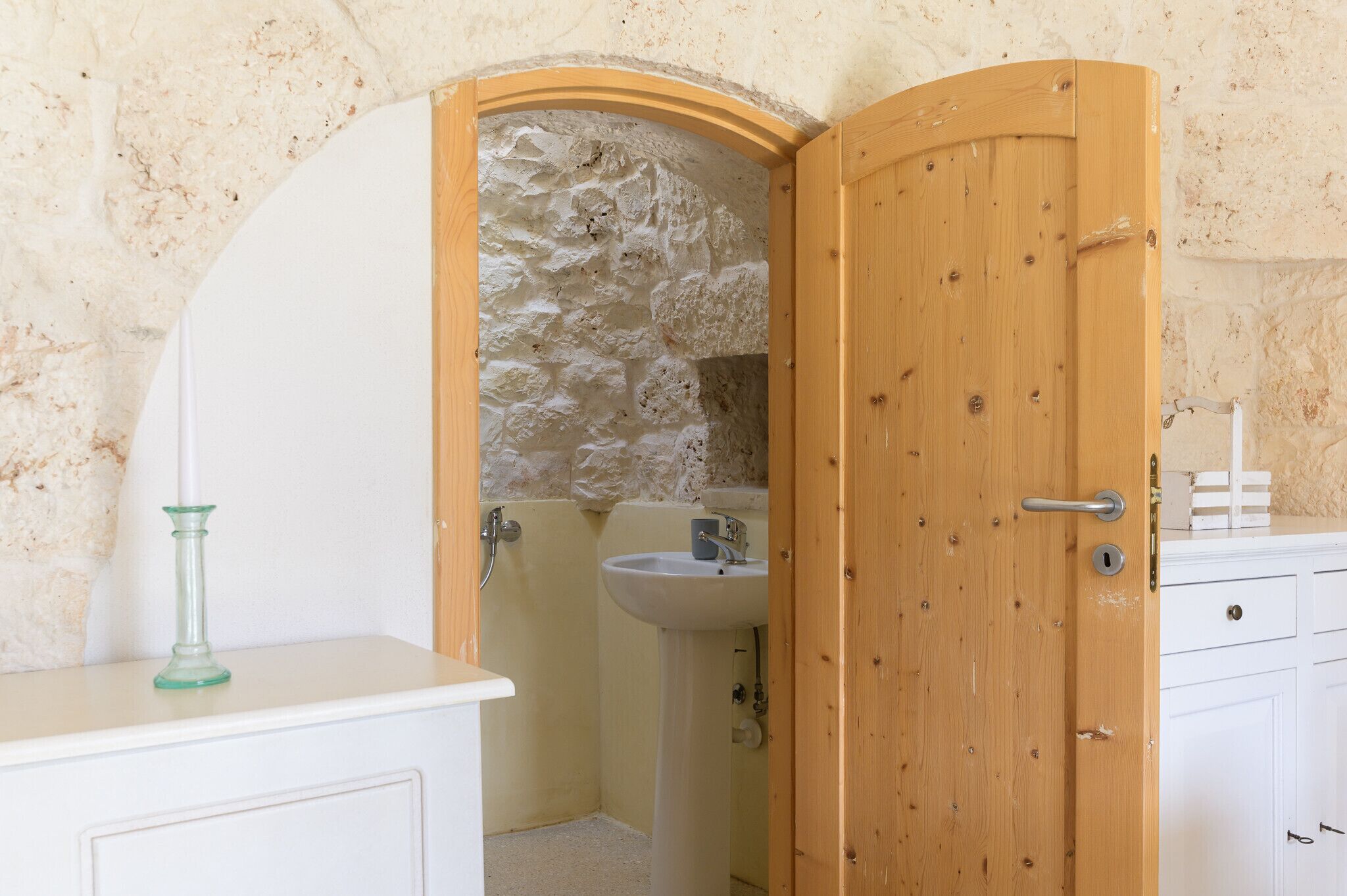A bathroom with a stone wall, a wooden door, a sink, and a glass candle on a white counter.