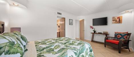A bedroom with a bed, a TV, a chair, and a door leading to another room.