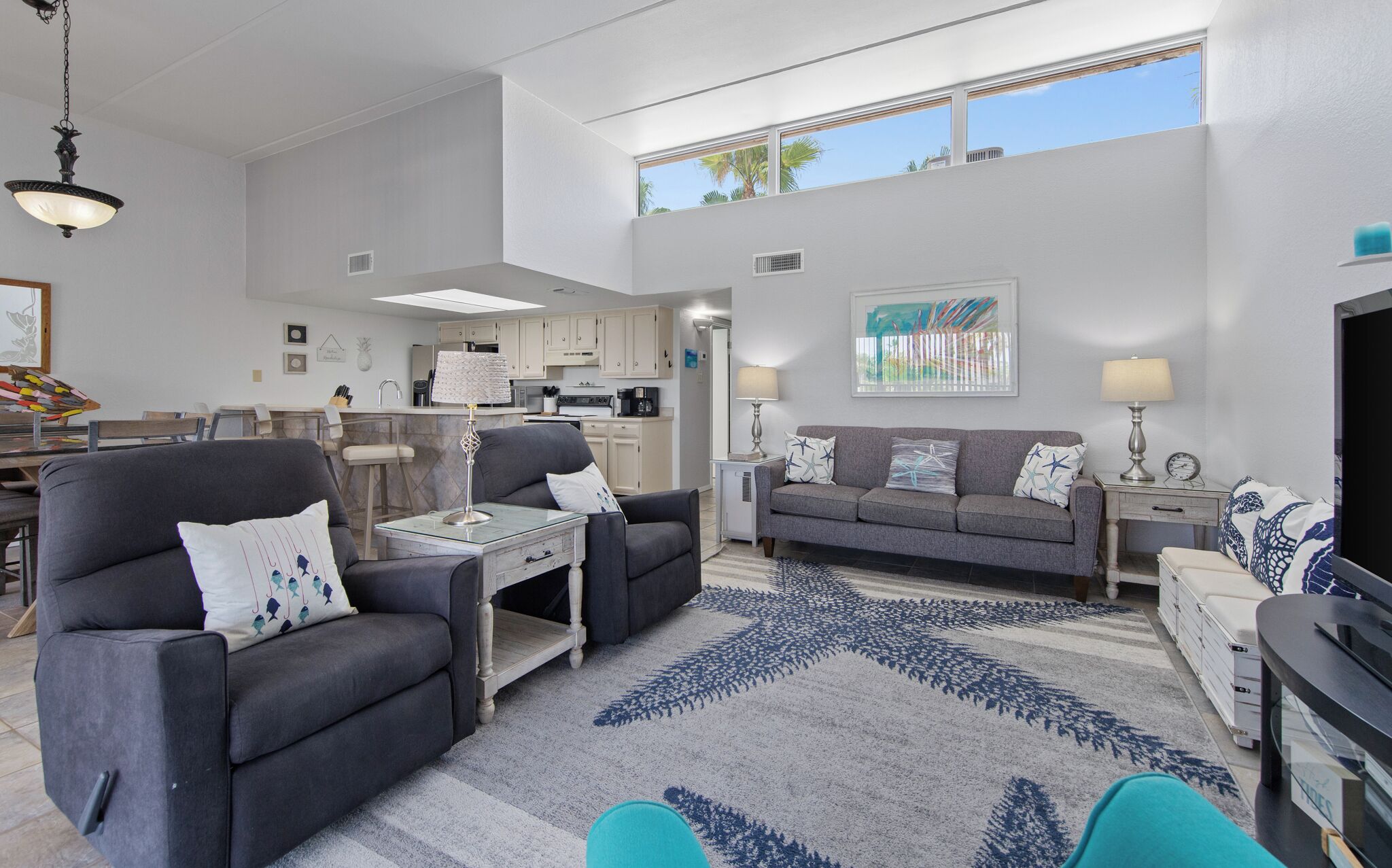 Una sala de estar moderna con un sofá gris, una mesa de centro y un mueble para televisión. Se ve una cocina al fondo con armarios blancos y una zona de comedor.