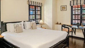 A hotel room with a large bed, a desk with a chair, and two windows with striped valances. - La Maison by Dia Hotels and Homes (Panaji)
