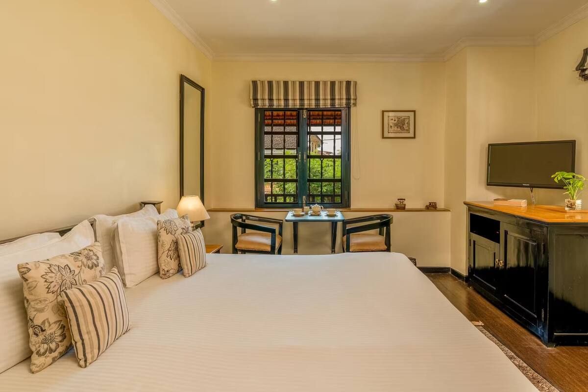 Ein Hotelzimmer mit einem großen Bett, einem Schreibtisch mit Stühlen, einem Fernseher und einem Fenster mit Blick auf Grünflächen.