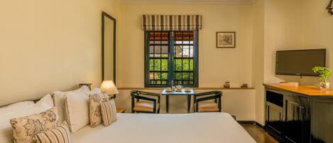 A hotel room with a large bed, a desk with chairs, a television, and a window with a view of greenery.