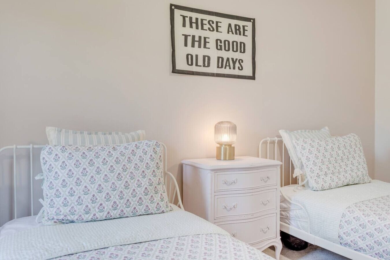 A bedroom with two beds, each with patterned bedding and pillows, a bedside table with a lamp, and a wall sign stating "These are the good old days."