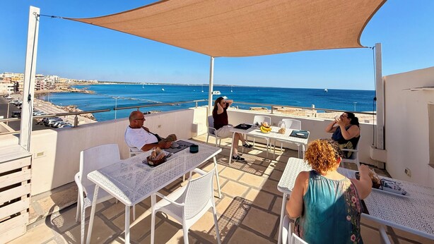 A terrace with white tables and chairs, a large shade sail, and a view of the sea and buildings. - Le Camere sul Golfo (Gallipoli)