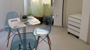 A small room with a table, chairs, a window with sheer curtains, a white cabinet, and a trash bin. - Le Camere sul Golfo (Gallipoli)