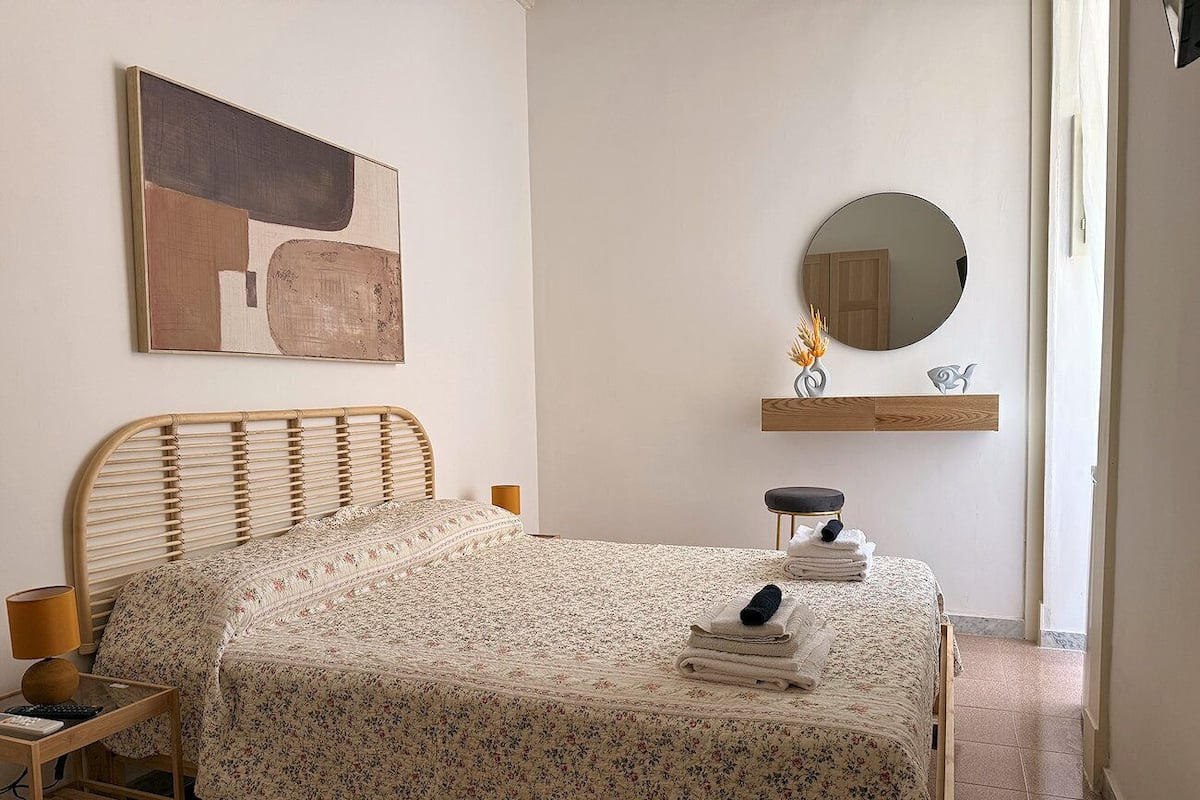 A bedroom with a bed, a bedside table, a lamp, a mirror, and a wall-mounted shelf with towels.